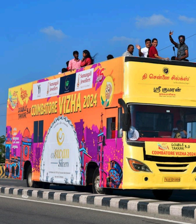 People riding on the open top of a yellow Coimbatore Vizha double-decker bus